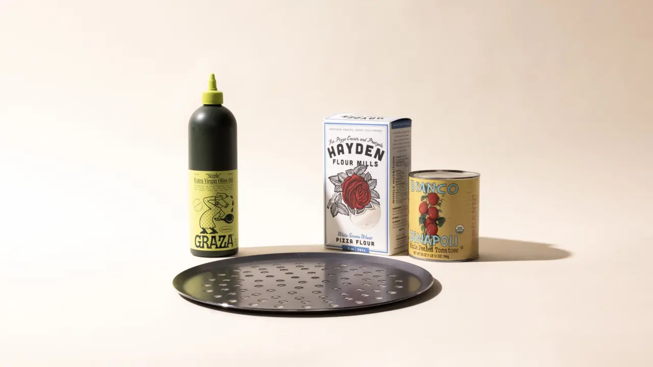 A bottle of olive oil, a packet of flour, a can of tomatoes, and a pizza tray arranged on a light background.