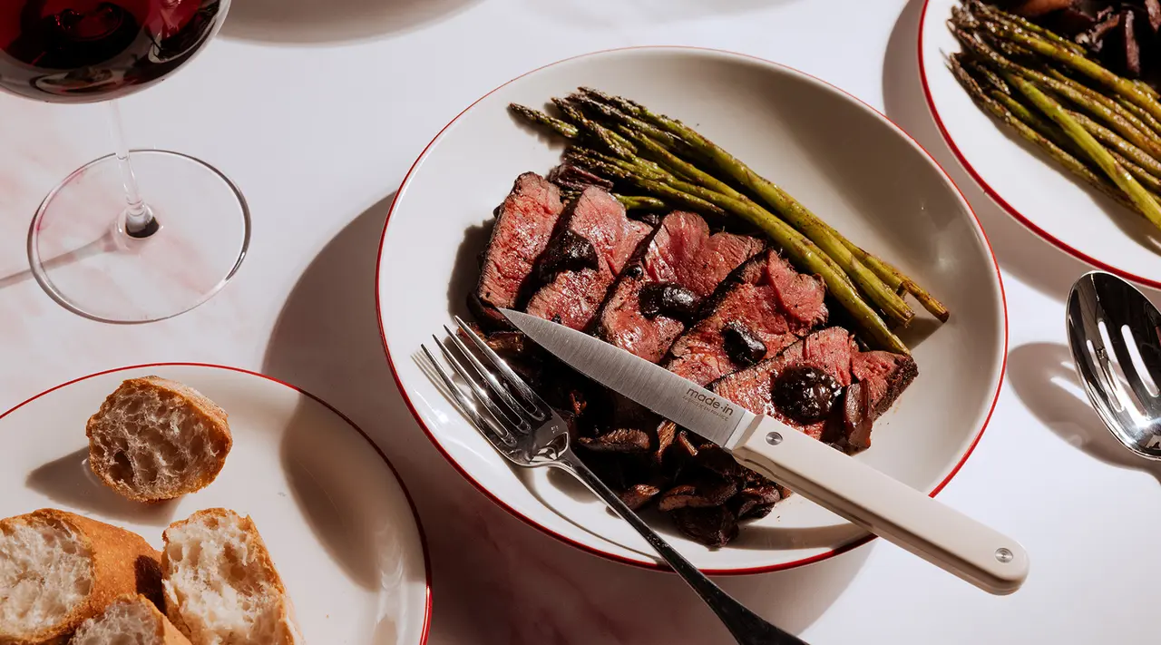 A well-prepared steak dinner with asparagus, slices of bread, and a glass of red wine is served on a table set with elegant dinnerware.