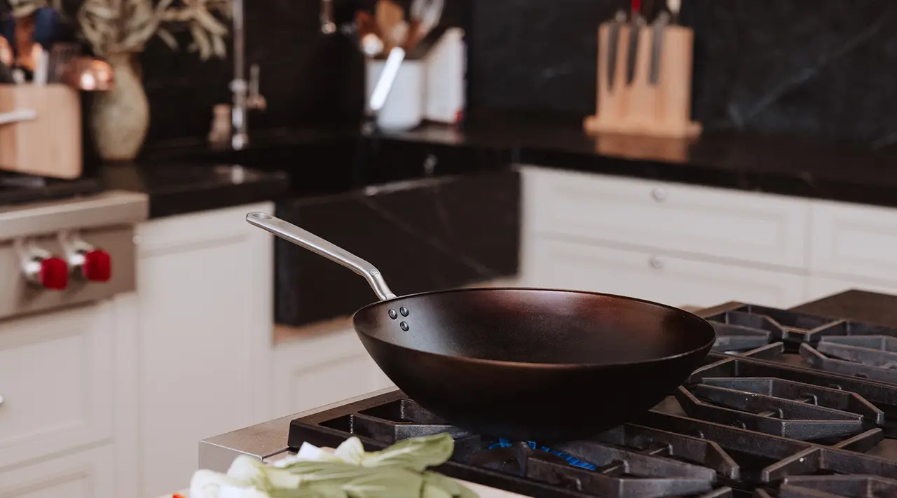 A black frying pan sits on a gas stovetop in a modern kitchen, with vegetables in the foreground.