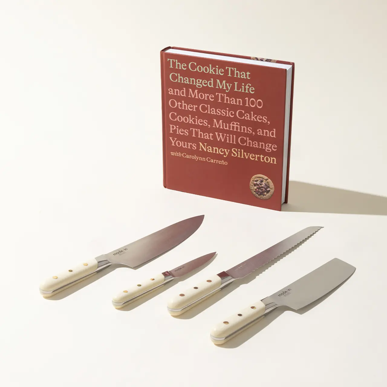 A cookbook titled "The Cookie That Changed My Life" is propped up in the background with a set of four kitchen knives displayed in the foreground on a light surface.