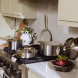 A modern kitchen scene features stainless steel cookware with gold handles, a vase of white flowers, and fresh vegetables on a countertop.