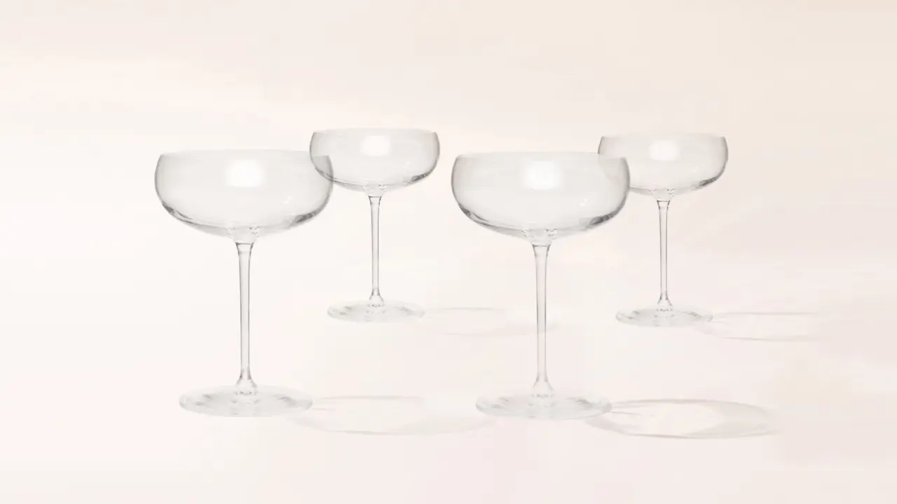 Four empty, clear coupe glasses are neatly lined up, casting soft shadows on a light surface.