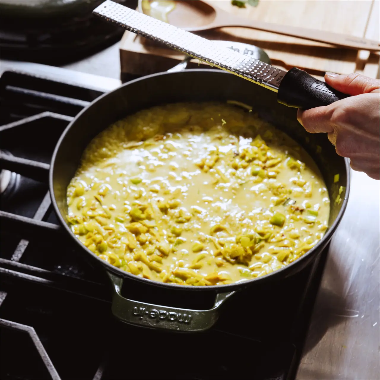 A hand is holding a grater over a pot of bubbling yellow soup or sauce on a stove.