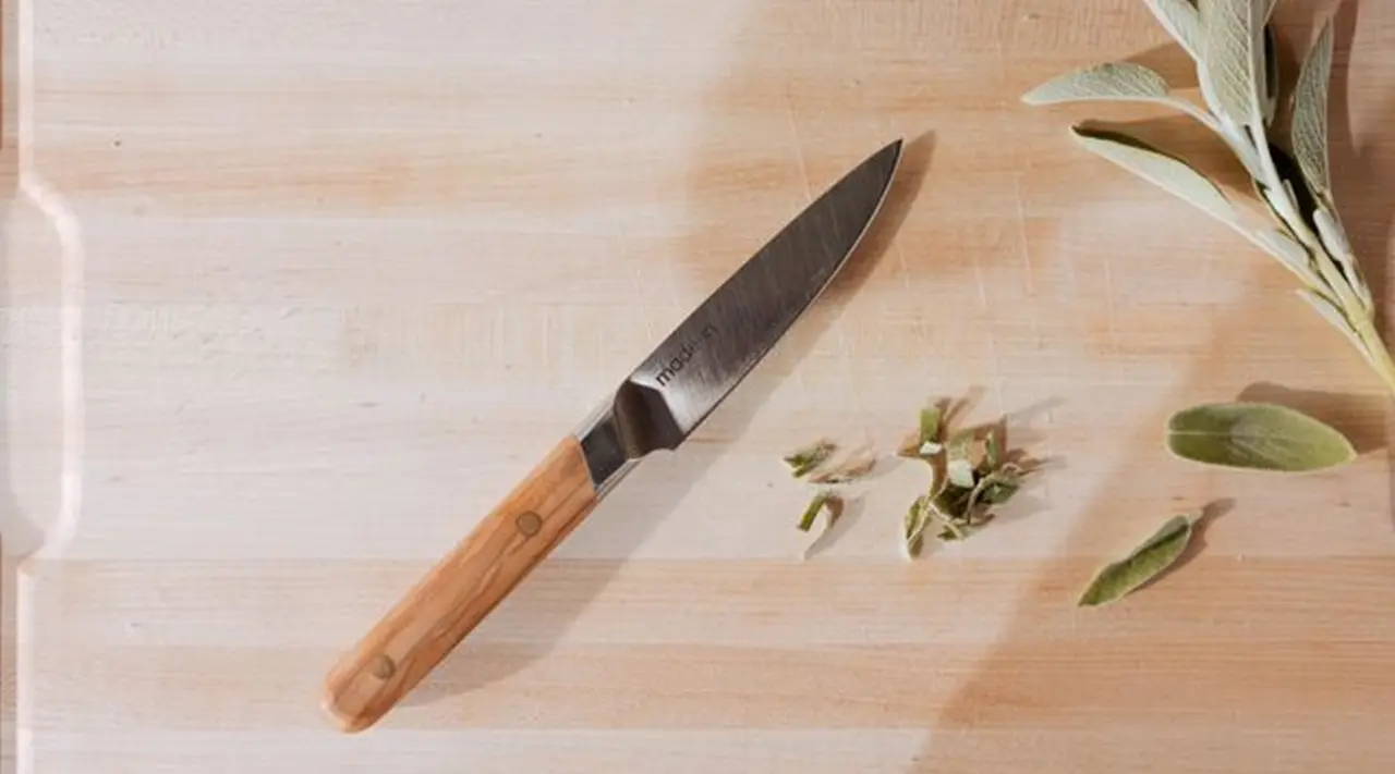 A kitchen knife lies on a chopping board next to some fresh green herbs.