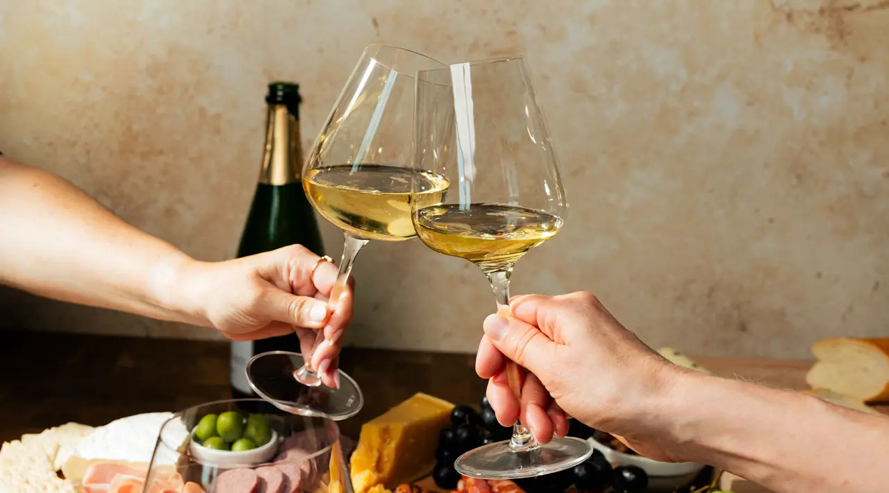 Two people are toasting with glasses of white wine over a table with various cheeses and snacks.