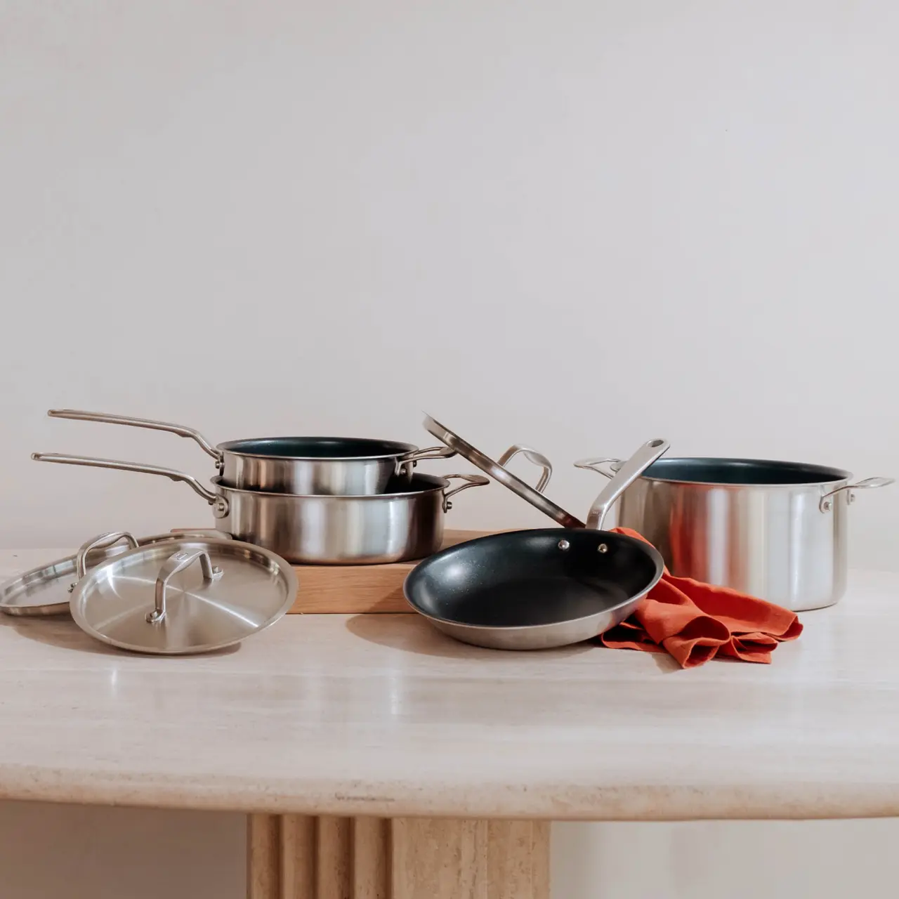 A set of stainless steel cookware including pots, a pan, and lids arranged neatly on a wooden table with a plain background.