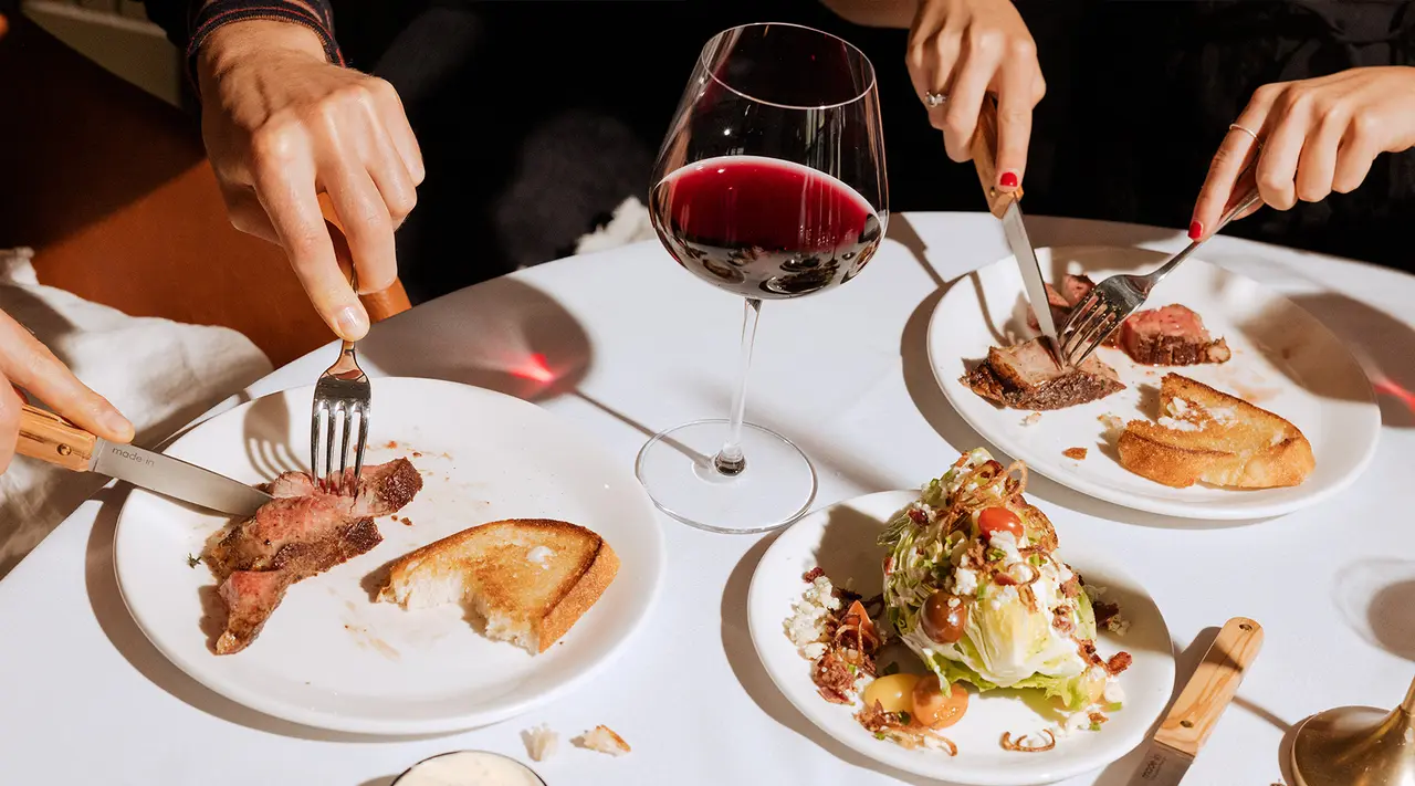 Diners enjoy a meal of steak and salad with a glass of red wine on an elegantly set table.