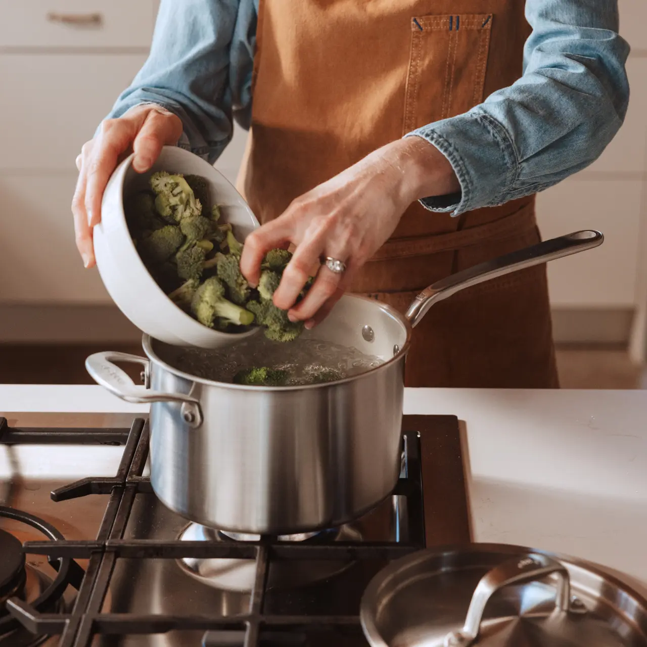 A person wearing an apron pours broccoli from a bowl into a pot of boiling water on a stove.