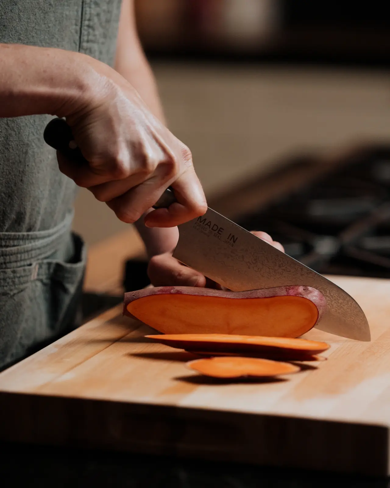 A person slices a sweet potato using a large kitchen knife on a wooden cutting board.