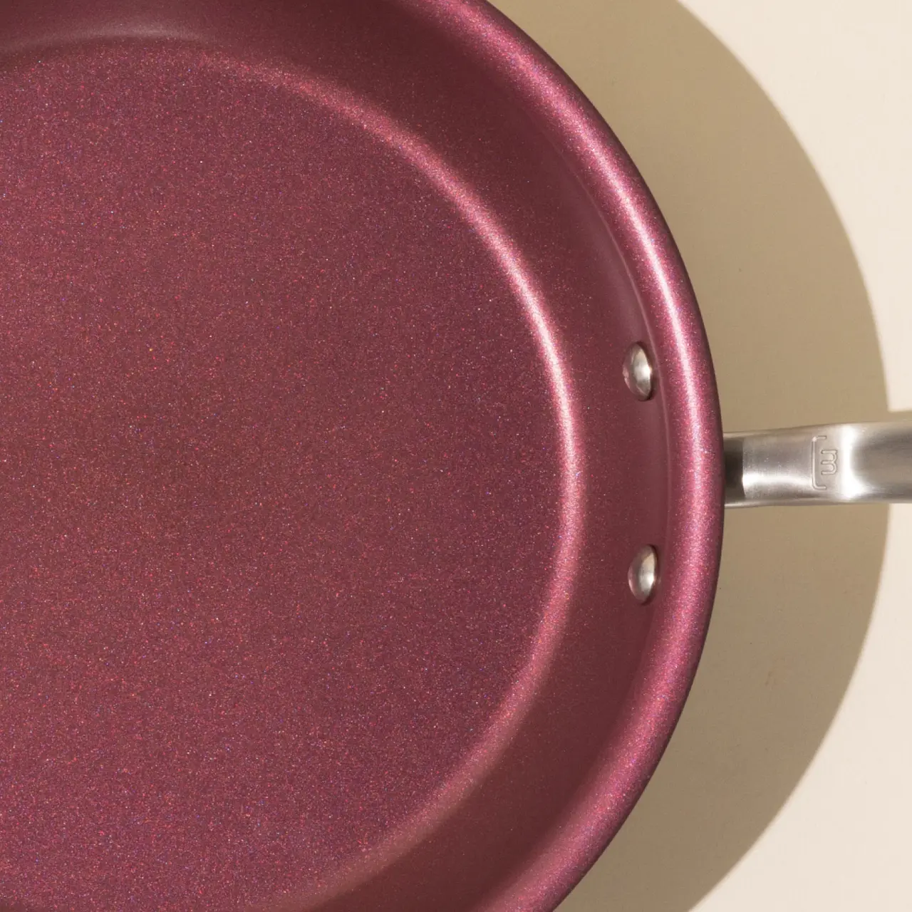 A close-up of a burgundy colored non-stick frying pan with a silver handle, casting a shadow on a beige surface.