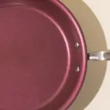 A close-up of a burgundy colored non-stick frying pan with a silver handle, casting a shadow on a beige surface.