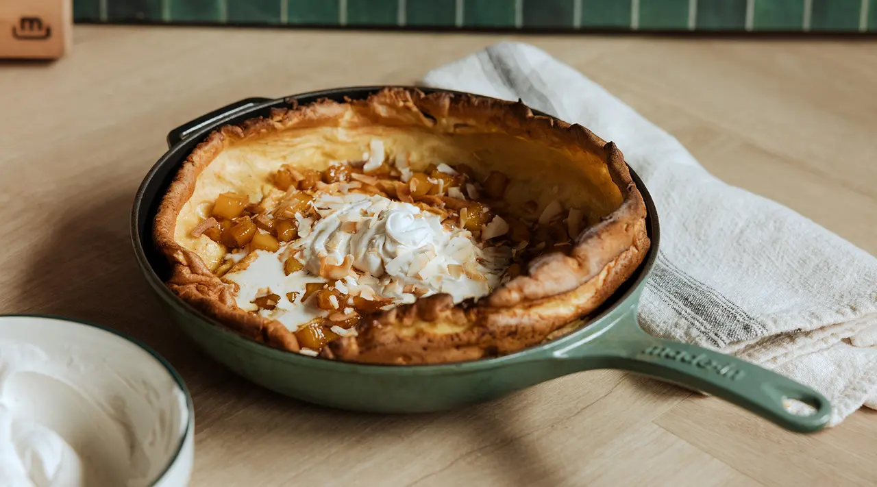 A freshly baked Dutch baby pancake is garnished with whipped cream and nuts, served in a green cast-iron skillet on a wooden table with a dollop of cream on the side.