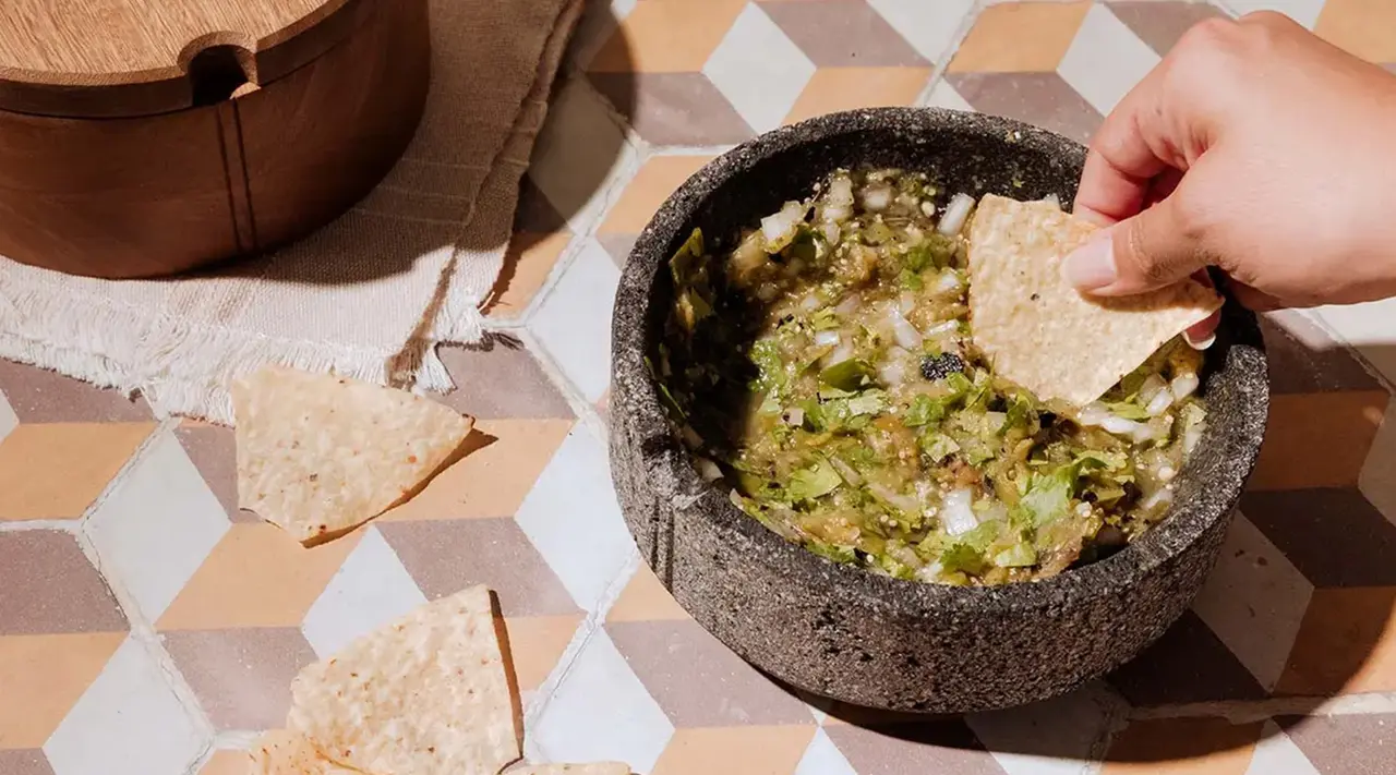 A person is dipping a tortilla chip into a bowl of salsa verde on a patterned surface, with a woven basket visible in the background.