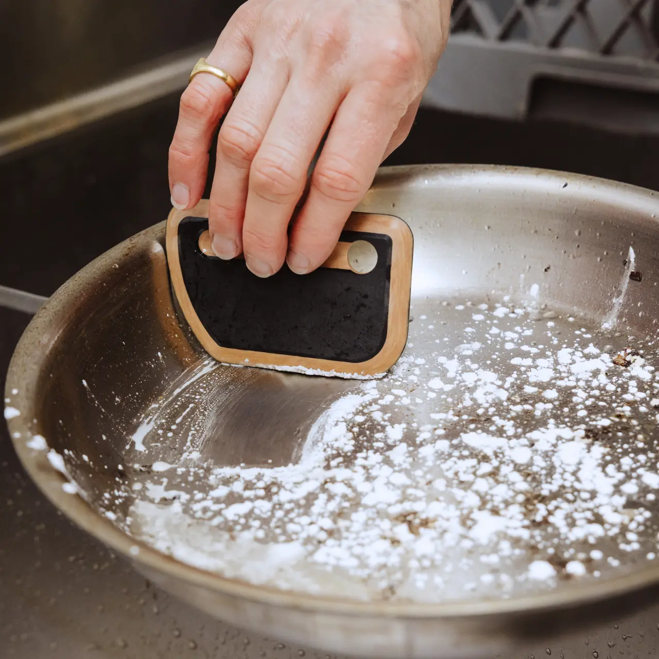 A hand holds a small cleaning tool over a stainless steel pan with some residue and powder on the bottom.