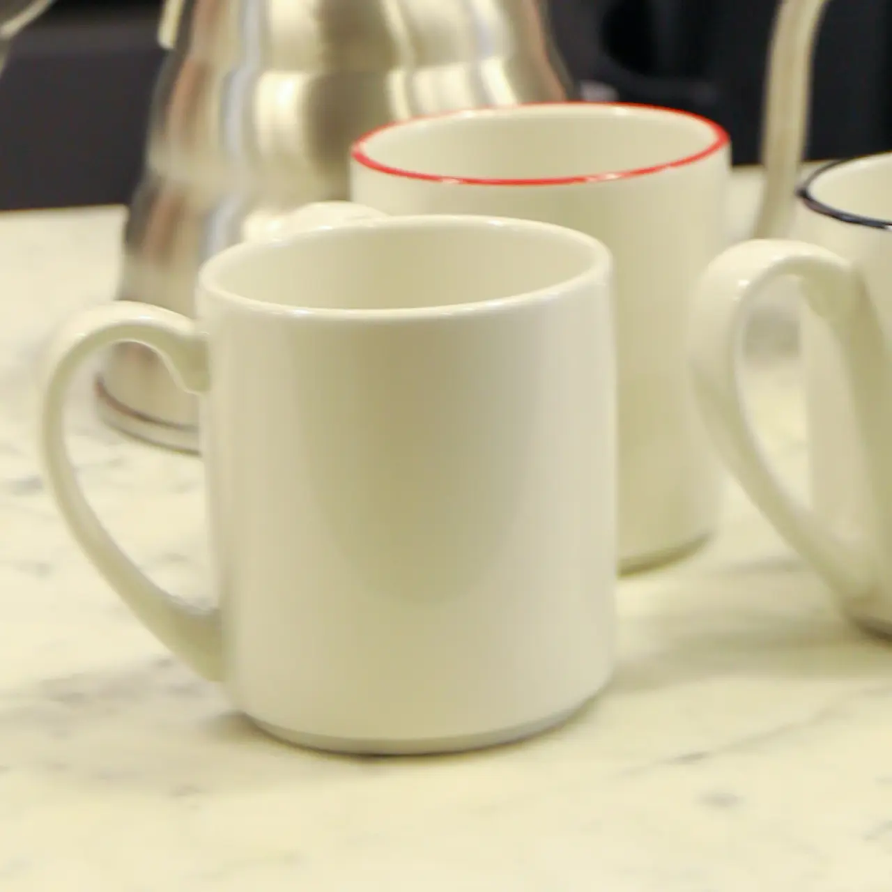 Three white mugs sit on a countertop with a stainless steel kettle in the background.
