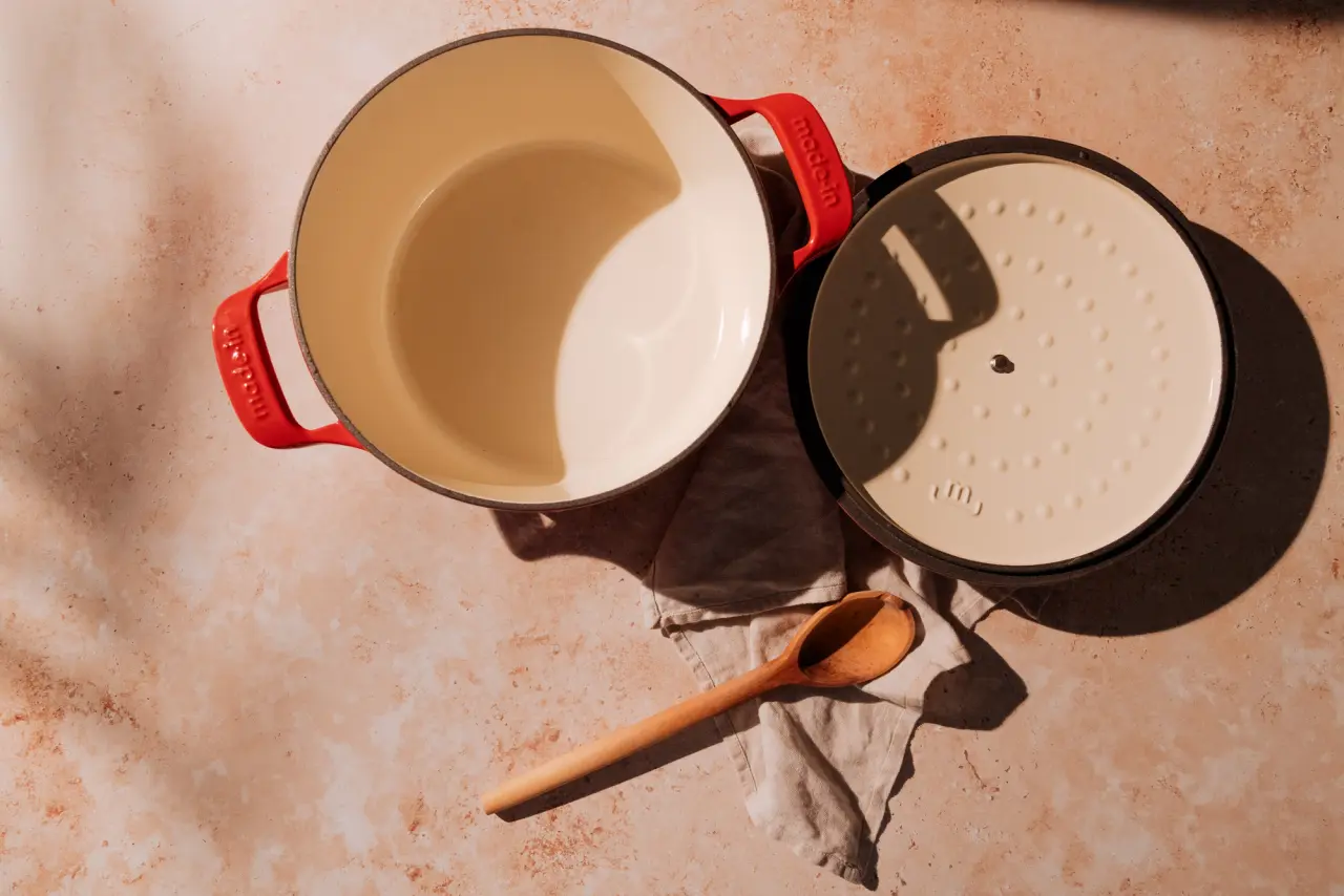 An enameled cast iron Dutch oven with the lid off and a wooden spoon beside it, on a textured surface with shadows indicating directional light.