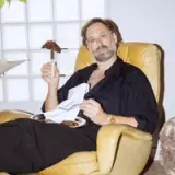 A man sits in a yellow chair, holding a plate of cookies in one hand and a cookie on a stick in the other, with a relaxed expression.