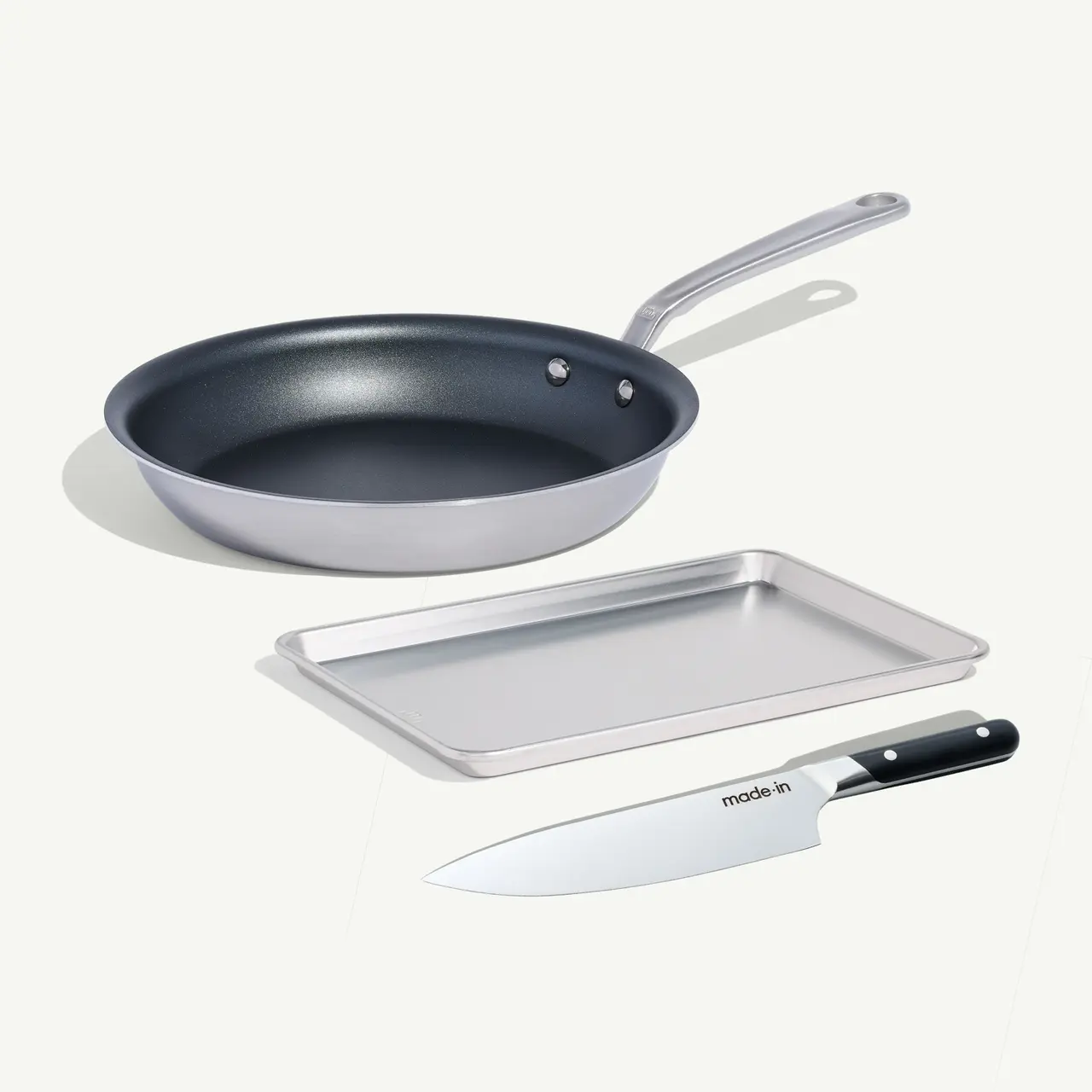 A frying pan, a baking tray, and a chef's knife are arranged neatly on a light background.