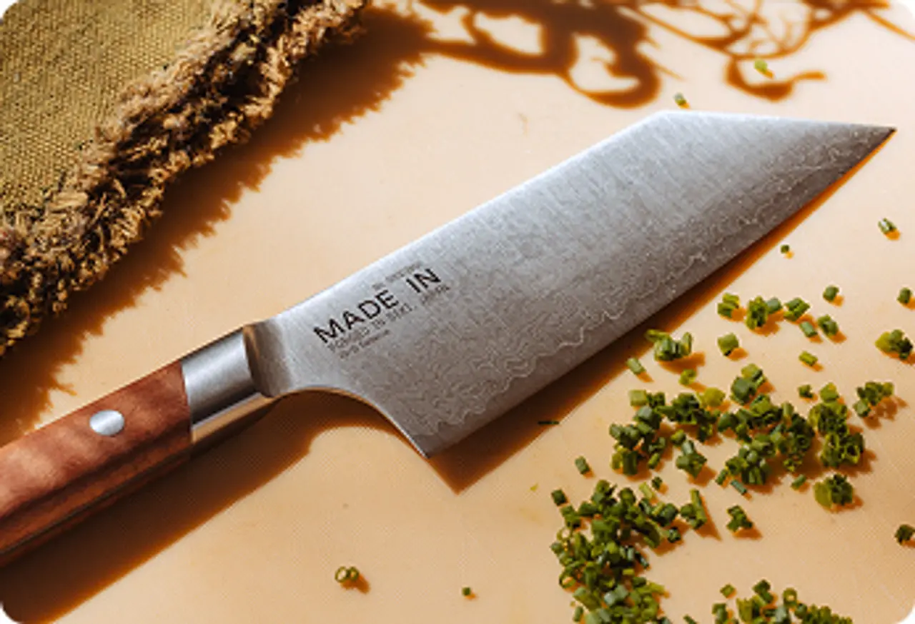 A sharp kitchen knife with a wooden handle rests on a light surface, surrounded by freshly chopped green herbs.