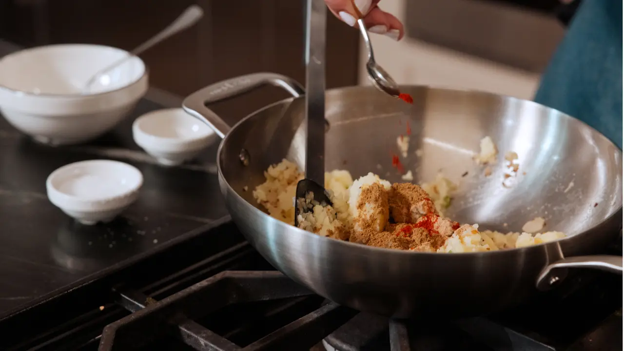 A person is stirring a mixture of rice and spices in a stainless steel pan on a stove, with bowls of ingredients nearby.