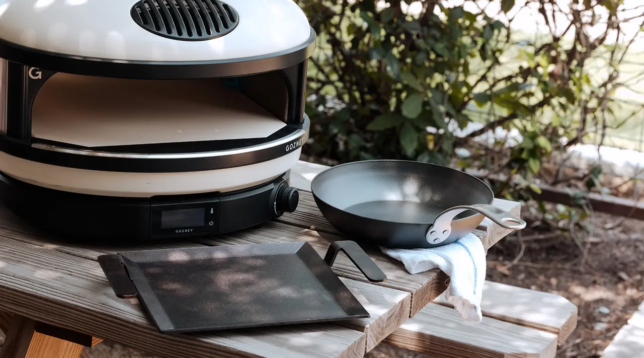 A portable pizza oven and a frying pan are set on a wooden table outdoors, suggesting an outdoor cooking setting.