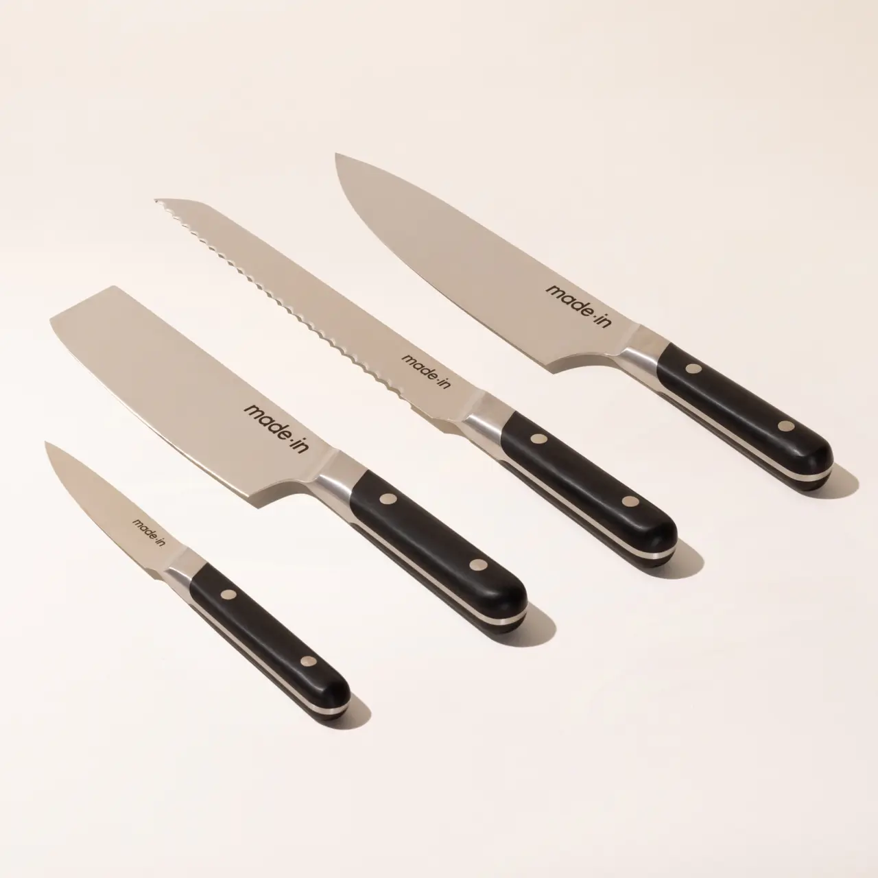 Four kitchen knives with black handles are displayed on a light background, consisting of a chef's knife, a serrated bread knife, a utility knife, and a paring knife.