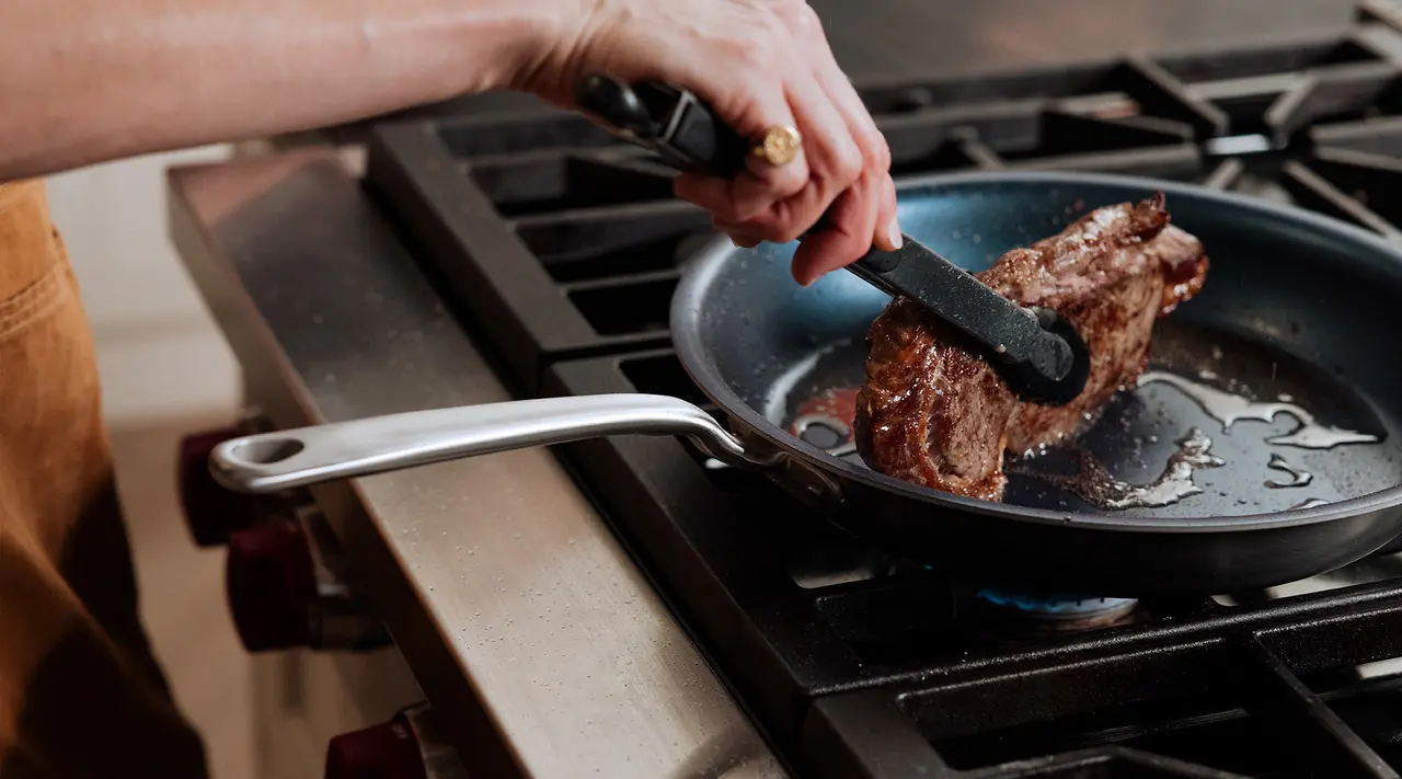 A person sears a steak in a pan on a stovetop, using tongs to handle the meat.