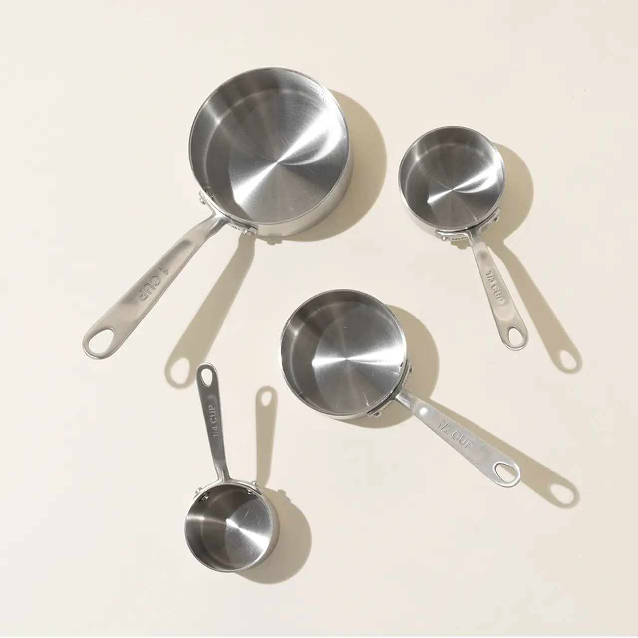 A set of four stainless steel measuring cups with different capacities are arranged on a light surface.