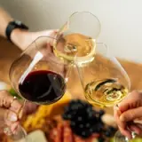Three wine glasses filled with red and white wine are clinking together in celebration, surrounded by a cheese and charcuterie spread.