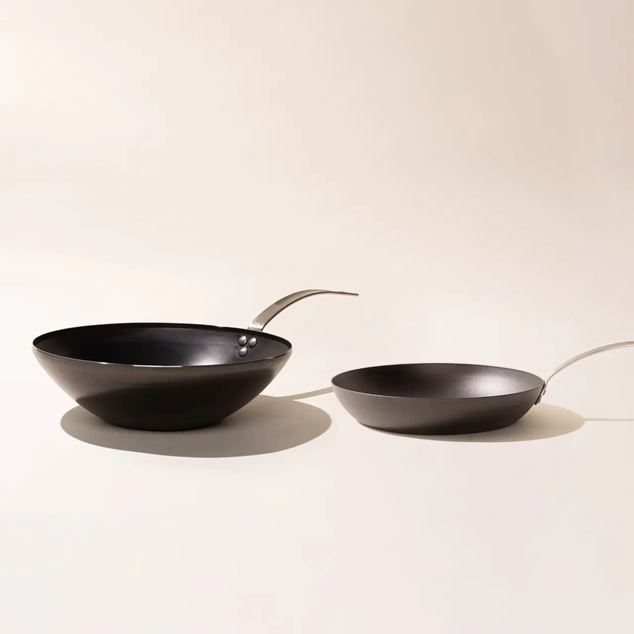 Two black frying pans with elongated, curved handles are positioned opposite each other, appearing to mimic a conversation or interaction, against a neutral background.