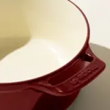 The image shows a close-up of a red enameled cast iron pot with a cream interior, focusing on the pouring spout with the brand name embossed.