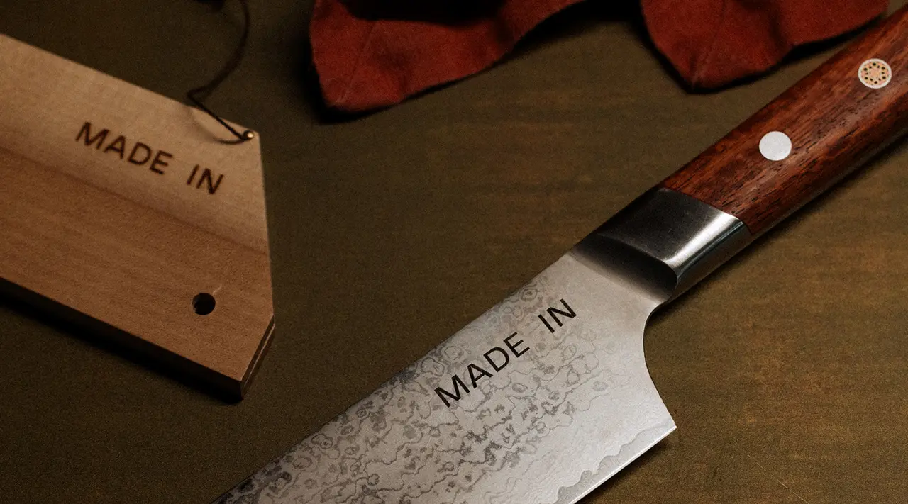 A knife with a wooden handle and "MADE IN" etched on the blade lies next to a similarly labeled tag on a textured surface.