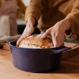 Hands are placing a freshly baked loaf of bread into a blue cooking pot on a wooden countertop.
