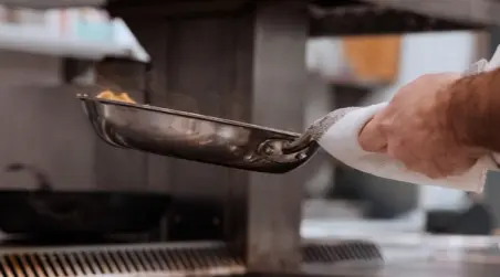 A hand holds a metal frying pan, skillfully tossing food while cooking in a professional kitchen.