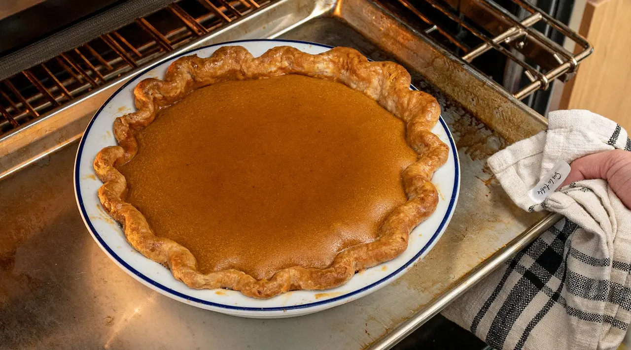 "A person is taking a freshly baked pumpkin pie out of the oven, holding the pie dish with a cloth to avoid getting burned."