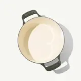 A round cooking pot with a glossy cream interior and dark green handles, featuring a heart-shaped design on the bottom.