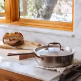 A stainless steel pot with a glass lid sits on a marble countertop next to a cutting board with sliced bread and a wooden spoon.