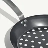 A metallic frying pan with a perforated surface and a curved handle is displayed against a light background.
