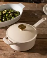 A cream-colored saucepan with a gold knob sits on a wooden table beside a dish of Brussels sprouts and a glass of red wine.