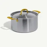 A stainless steel cooking pot with yellow handles and a matching lid sits on a light background.
