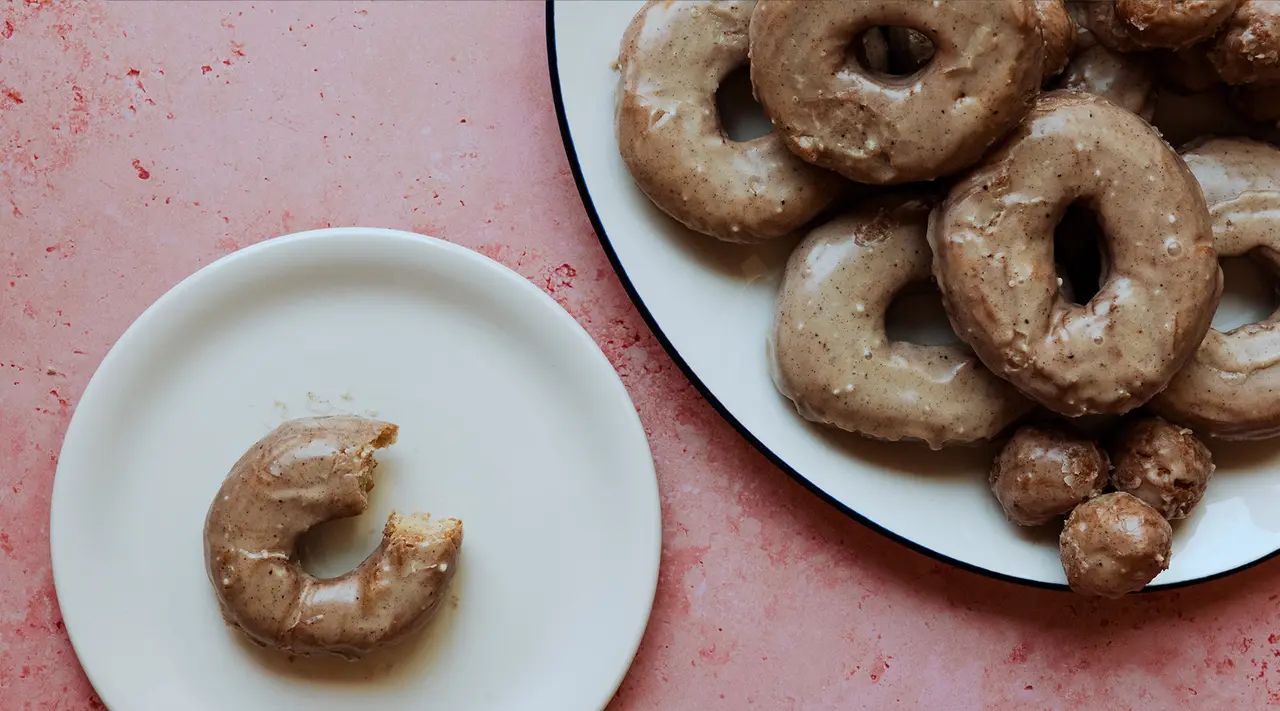 A plate with several whole glazed donuts and one partially eaten donut on a small plate to its left, all set on a table with a pink surface.