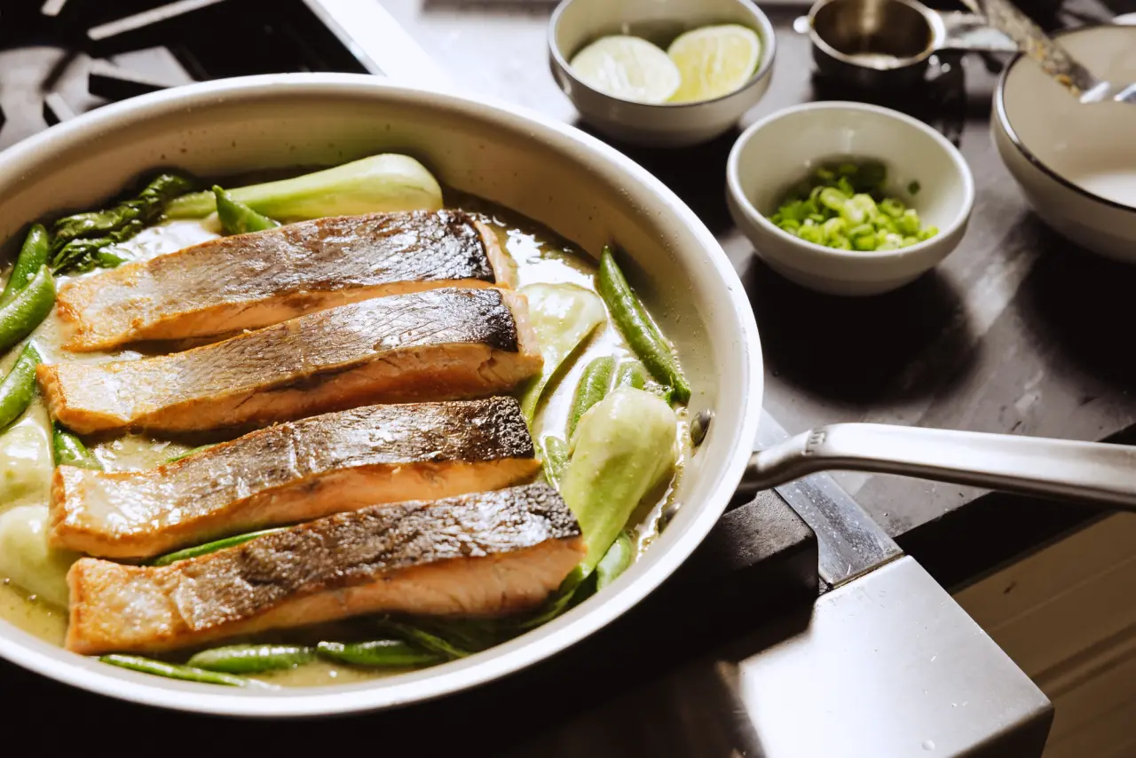 Four pieces of grilled salmon rest on a bed of bok choy and green beans in a skillet, accompanied by bowls of lime and sliced green onions.