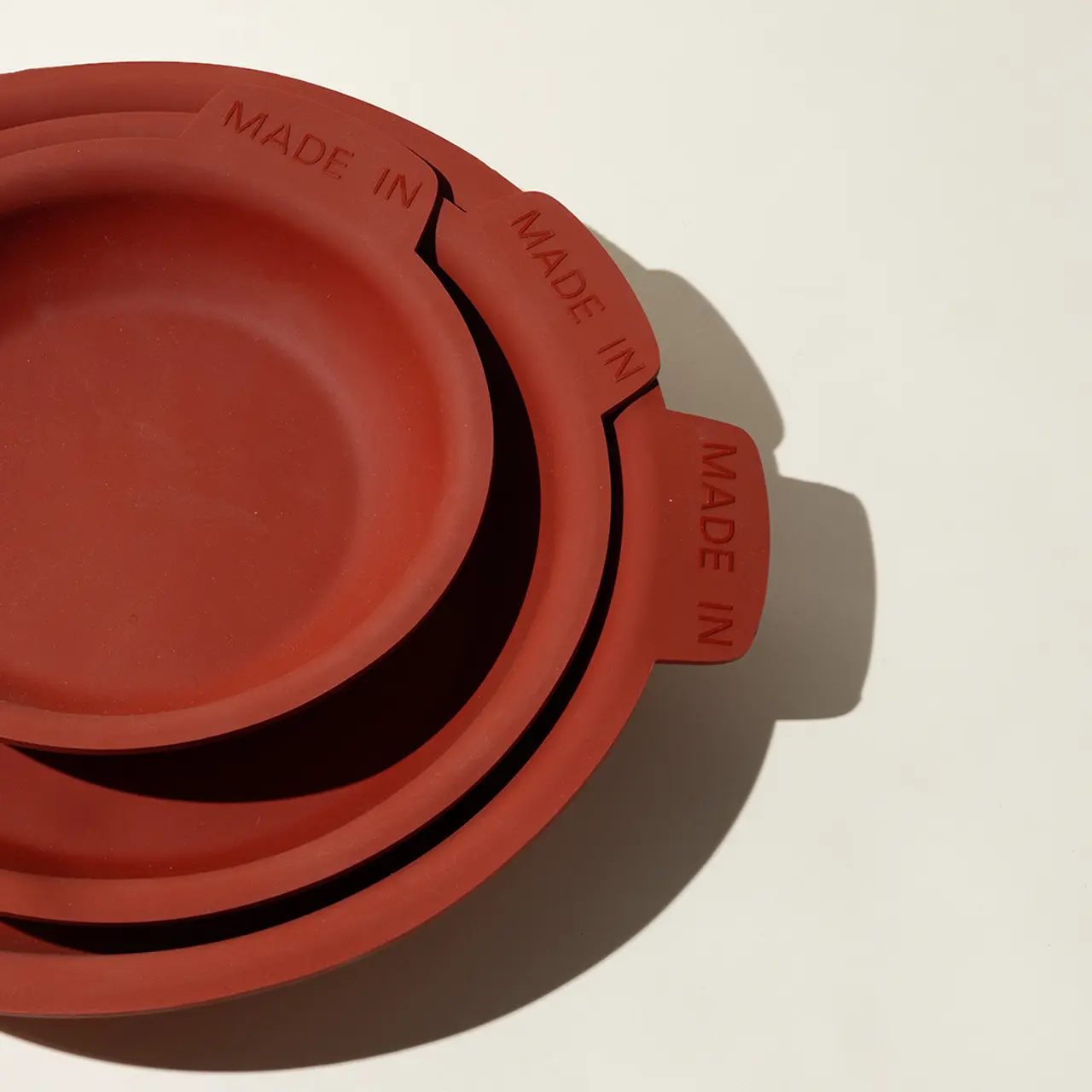 A stack of red ceramic plates of varying sizes with "MADE IN" embossed on their rims, casting shadows on a light surface.