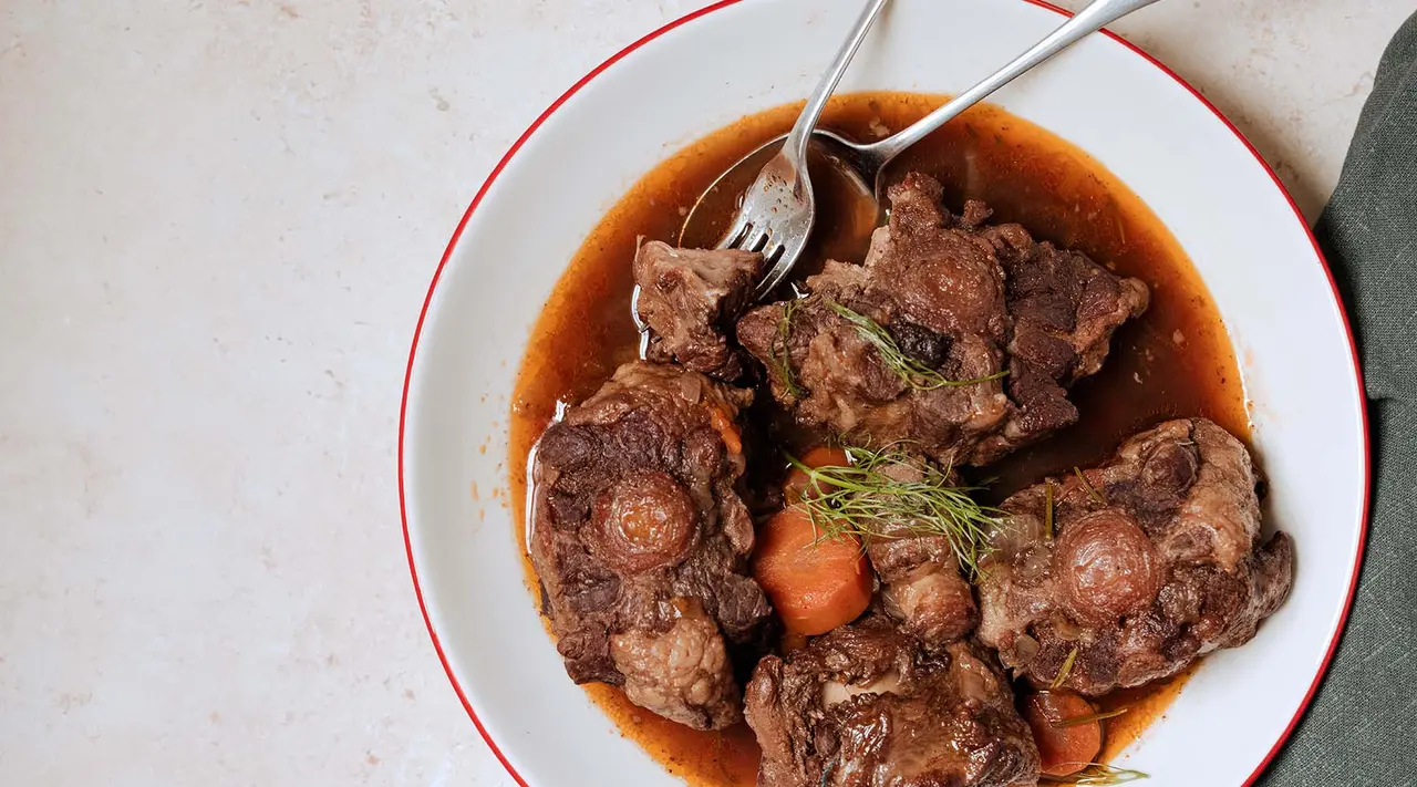 A bowl of beef stew with carrots and a sprig of herbs, accompanied by a fork on a light background.