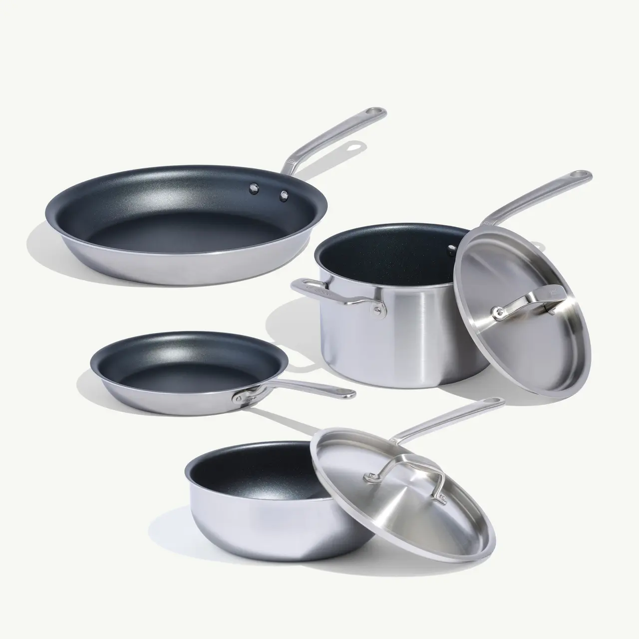 Assorted stainless steel cookware, including a frying pan, a saucepan with a lid, and smaller pots, arranged artfully.