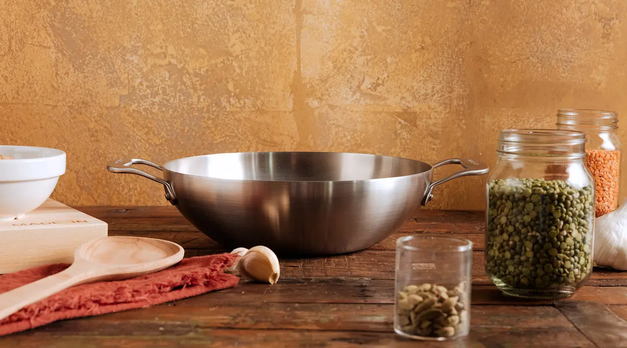 A stainless steel wok is centered on a kitchen counter surrounded by ingredients including lentils, seeds, garlic, and a cutting board with raw meat.