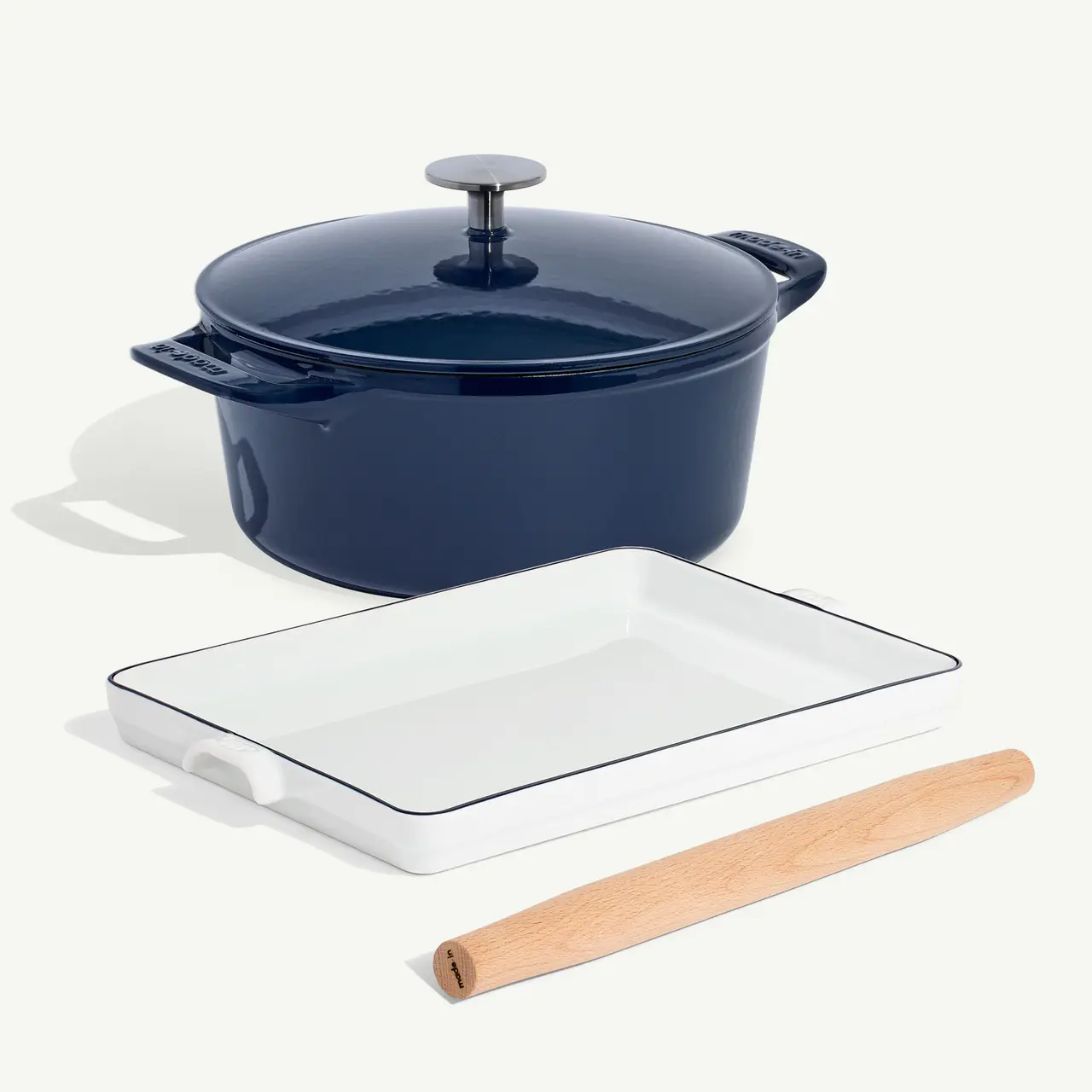 A navy blue dutch oven, a white rectangular baking dish, and a wooden rolling pin are neatly arranged on a light surface.