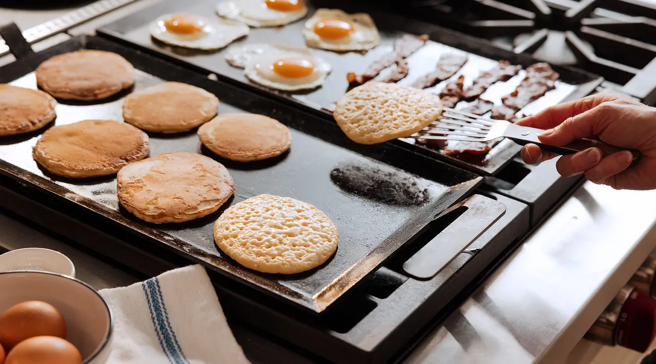 A variety of breakfast items, including pancakes, eggs, and bacon, are being cooked on a large stovetop griddle, with someone flipping a pancake.