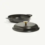 A black, cast iron frying pan with a matching lid sits on a light background, showcasing its sleek design and round shape.