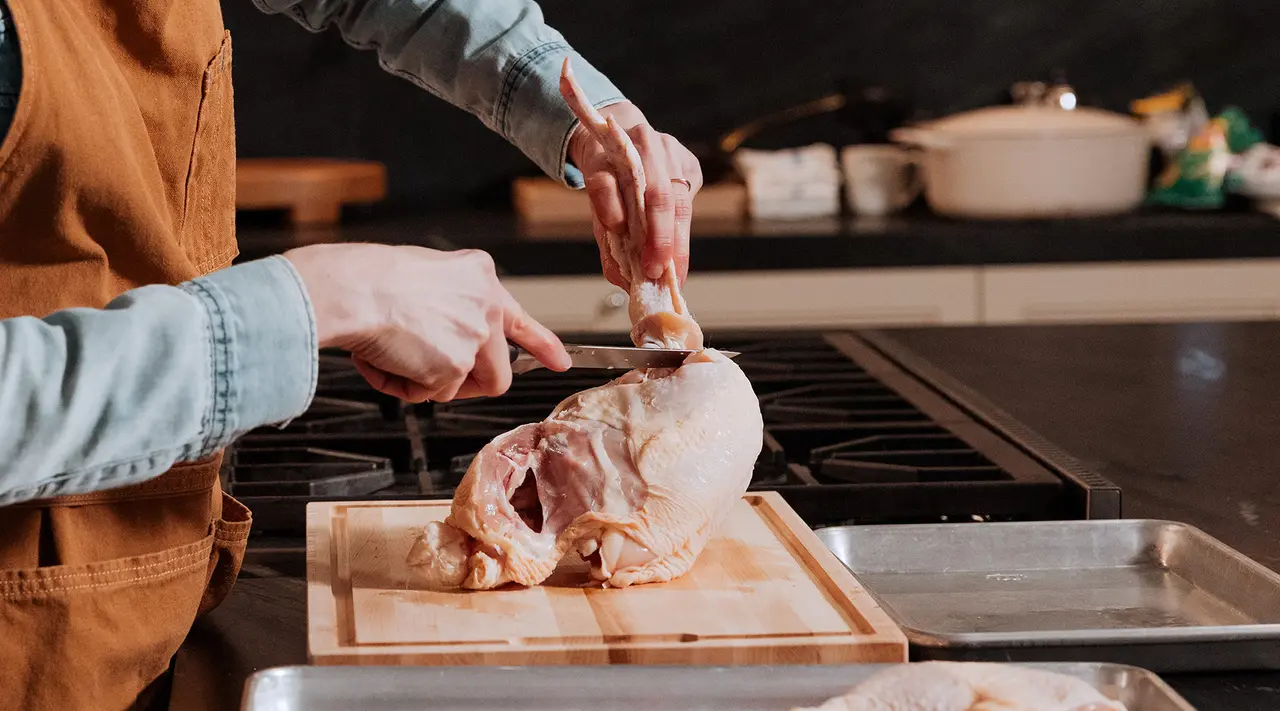 A person wearing an apron is preparing a raw chicken on a wooden cutting board in a kitchen.